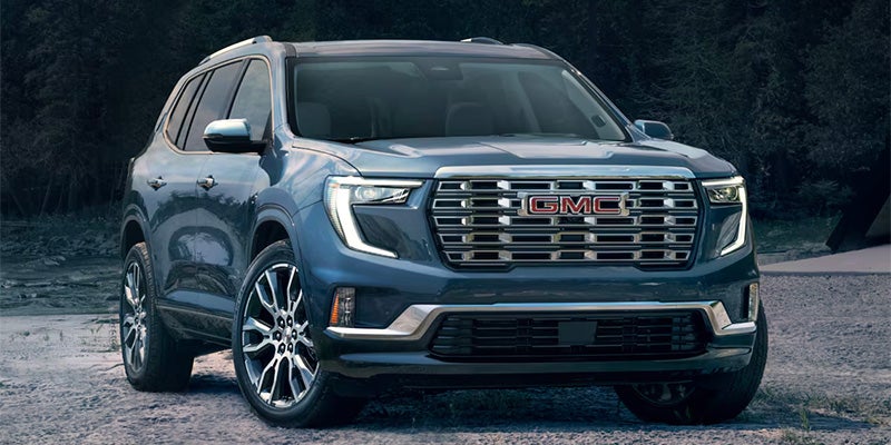 GMC SUV front view with bold chrome grille, parked on gravel near wooded background.